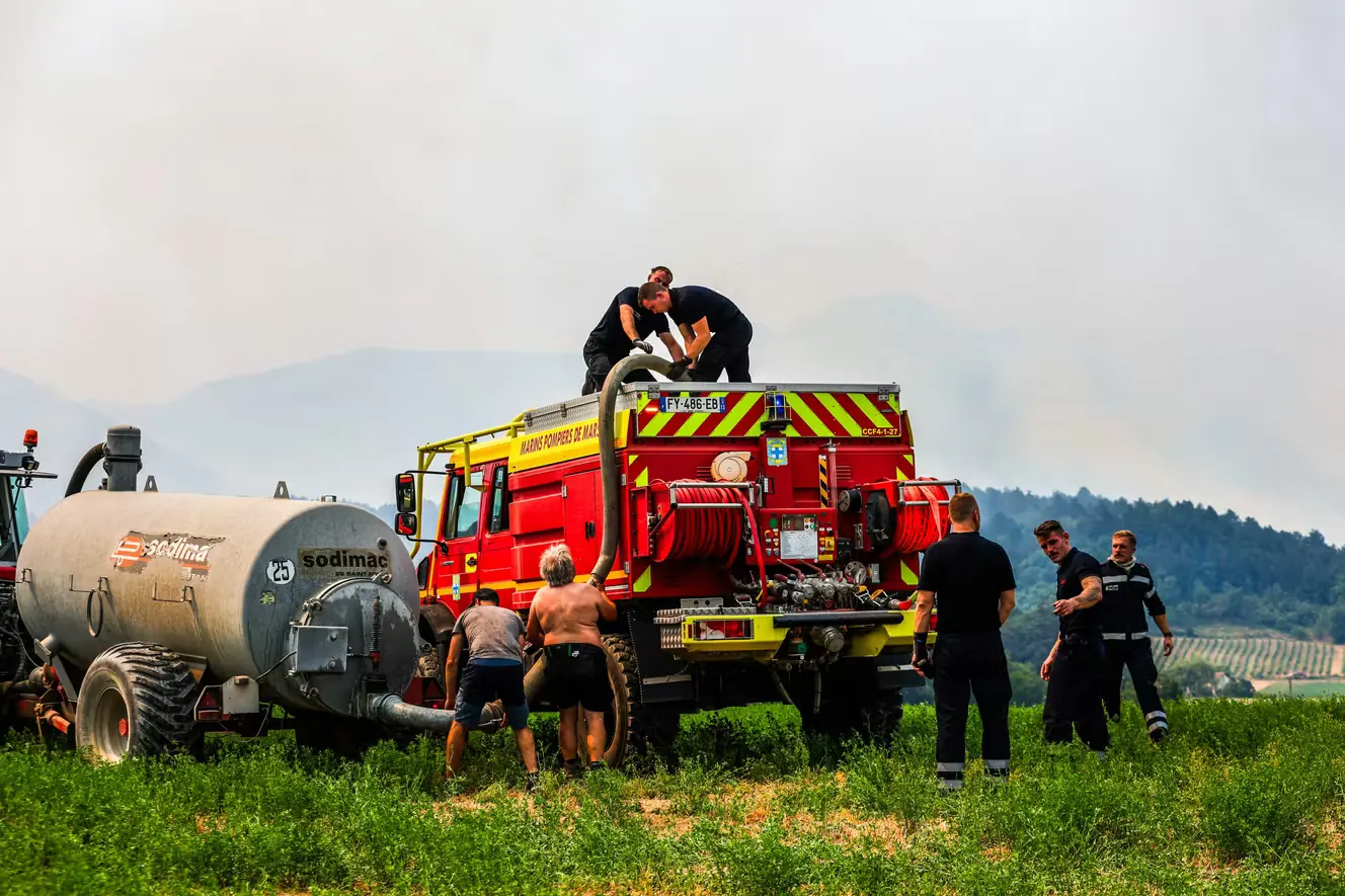 Dans un champ sous un ciel chargé de fumée, une citerne agricole remplit un camion de pompiers ; pompiers en tenue et habitants en short travaillent côte à côte.