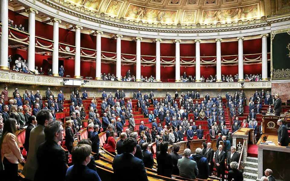 Hémicycle de l'Assemblée nationale : députés debout à leurs bancs et public dans les tribunes, lors d'une minute de silence.