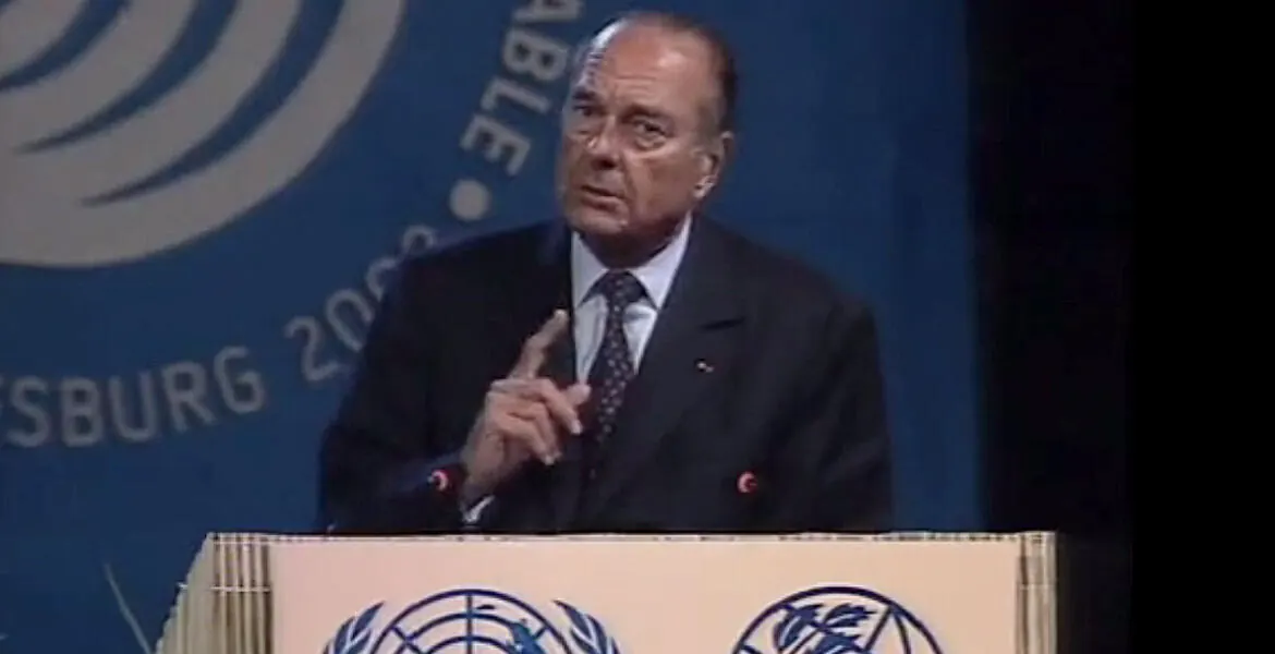 Un homme âgé en costume sombre, index levé, parle à un pupitre portant l'emblème des Nations unies, devant un fond bleu où se lit « ...sburg 200... ».