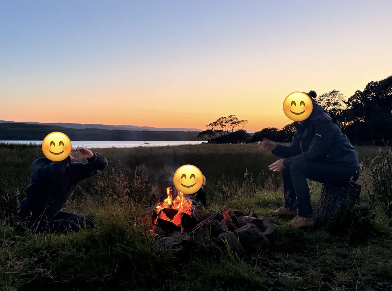 Trois personnes, visages masqués par des émojis, autour d'un petit feu de camp dans l'herbe, au bord d'un loch, au coucher du soleil.
