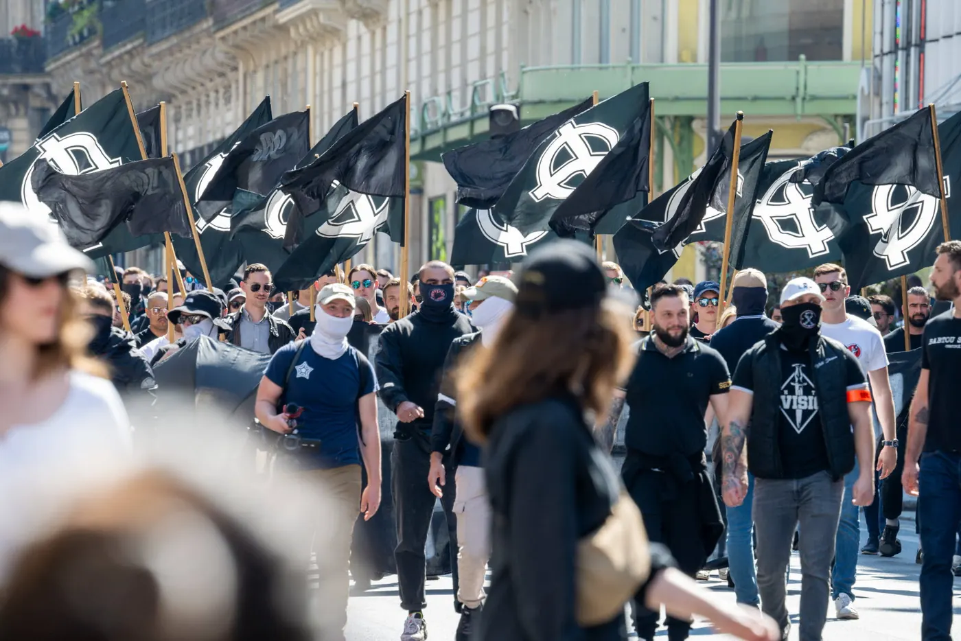 Défilé dans une rue de Paris : des dizaines de manifestants, plusieurs le visage masqué, marchent sous des drapeaux noirs frappés de croix celtiques blanches.