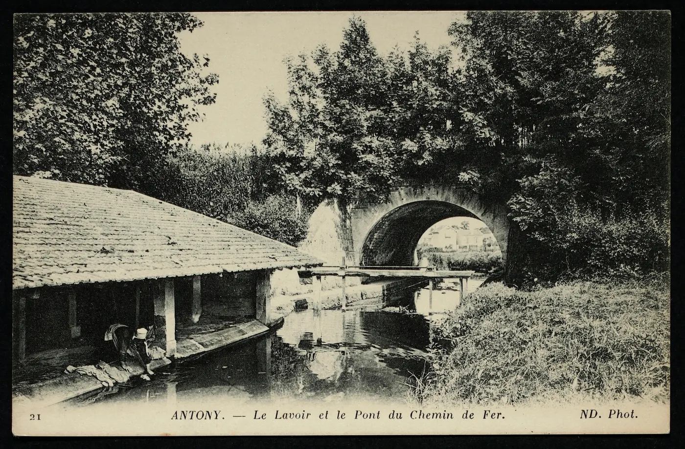 Carte postale ancienne « Antony, le lavoir et le pont du chemin de fer » : une laveuse s'active sous l'auvent d'un lavoir au bord d'un ruisseau qui passe sous l'arche du pont.