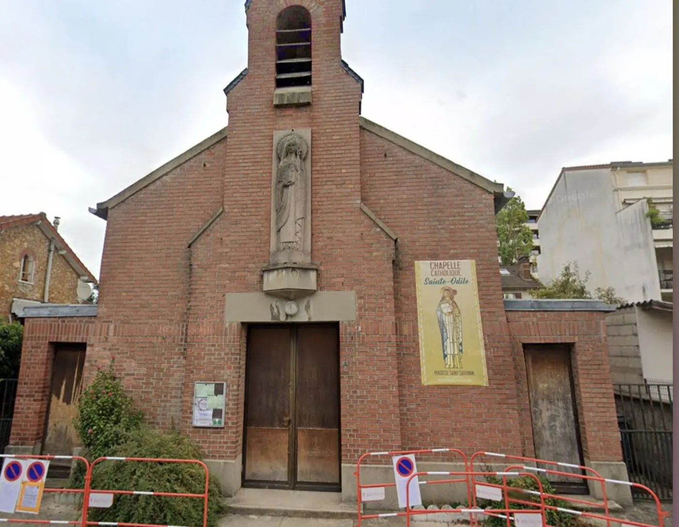 Vue actuelle de la chapelle catholique Sainte-Odile : façade de briques avec clocheton, statue sculptée au-dessus de la porte et bannière de la paroisse, derrière des barrières de chantier.