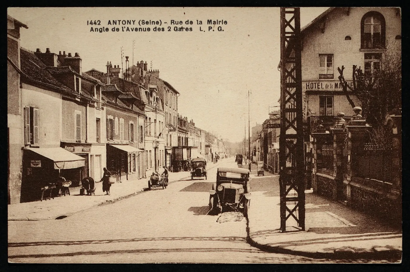Carte postale ancienne « Antony (Seine), rue de la Mairie, angle de l'avenue des 2 Gares » : rue bordée de commerces, deux automobiles anciennes et l'Hôtel de la Mairie à droite.