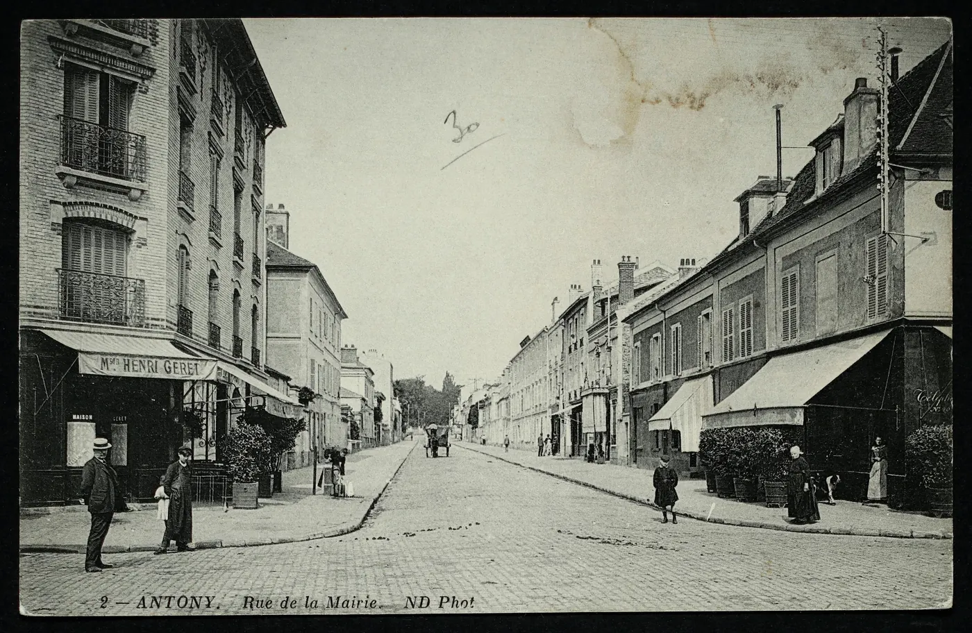 Carte postale ancienne « Antony, rue de la Mairie » : rue pavée bordée d'immeubles et de boutiques à auvents, avec des passants et une charrette au loin.