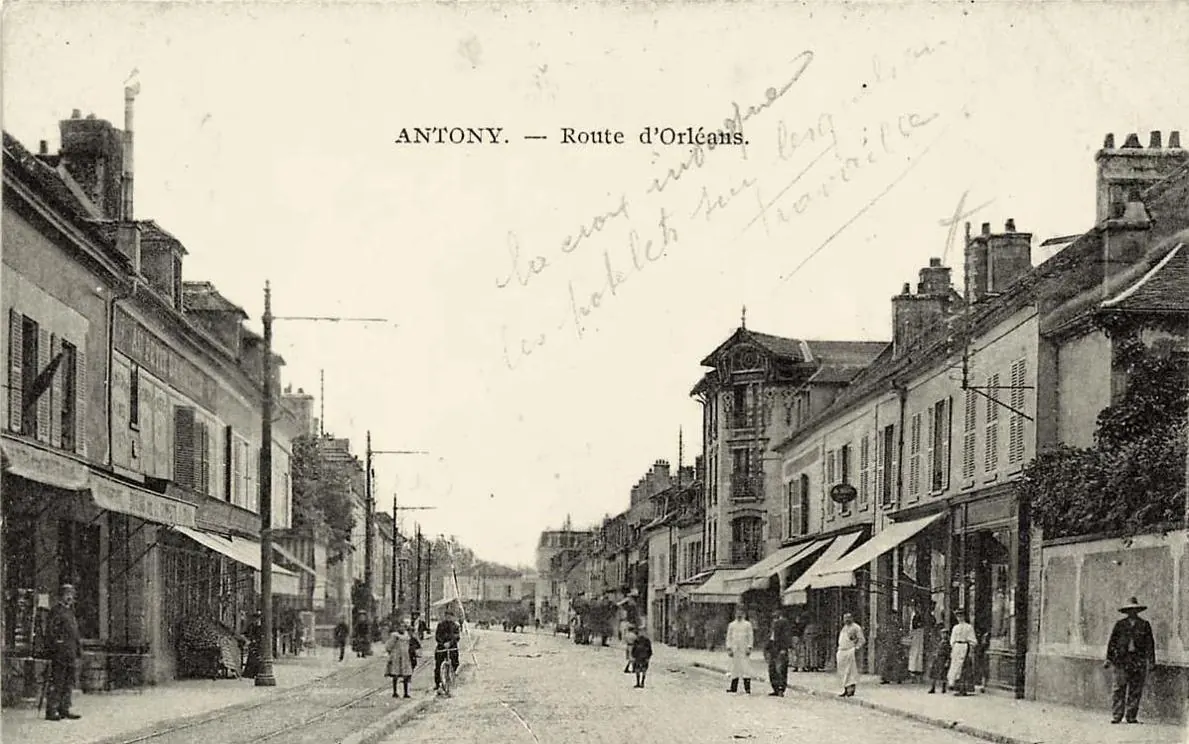 Carte postale ancienne « Antony, route d'Orléans » : rue animée avec rails de tramway, poteaux électriques, commerces à auvents et passants, avec un mot manuscrit en travers du ciel.