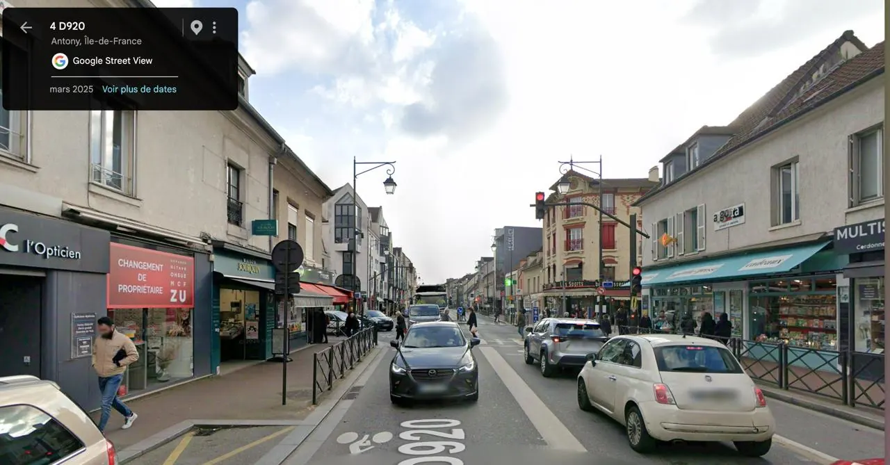 Vue Street View de mars 2025 de la D920 à Antony : la même rue, aujourd'hui bordée de commerces modernes, avec voitures, feux tricolores et réverbères.