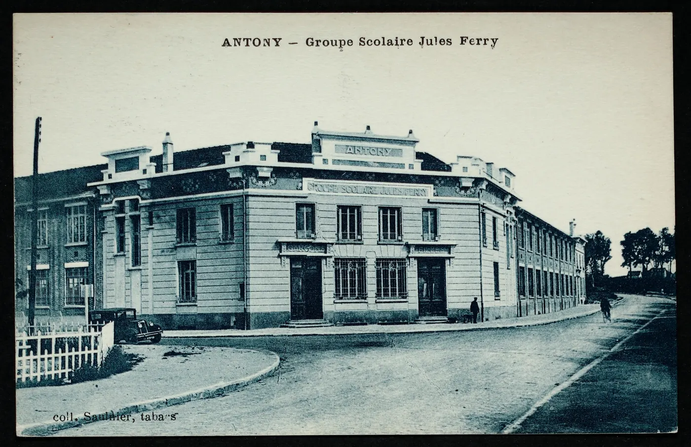 Carte postale ancienne « Antony, groupe scolaire Jules Ferry » : le même bâtiment d'angle, avec une automobile ancienne garée et un cycliste sur la route.