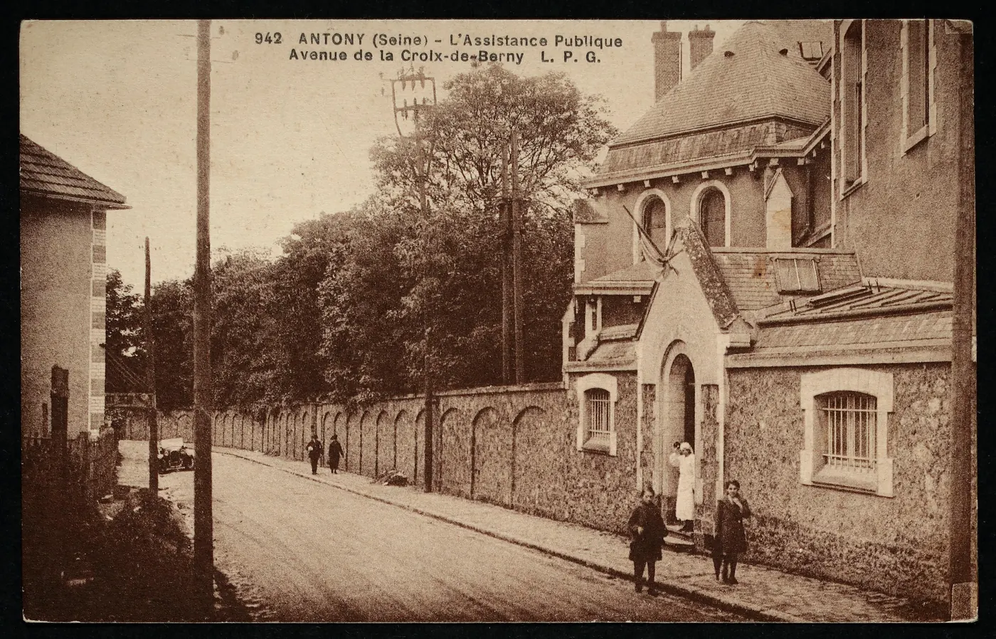 Carte postale ancienne « Antony (Seine), l'Assistance publique, avenue de la Croix-de-Berny » : un long mur de pierre et un bâtiment à porche, avec des enfants et une femme en tablier blanc sur le trottoir.