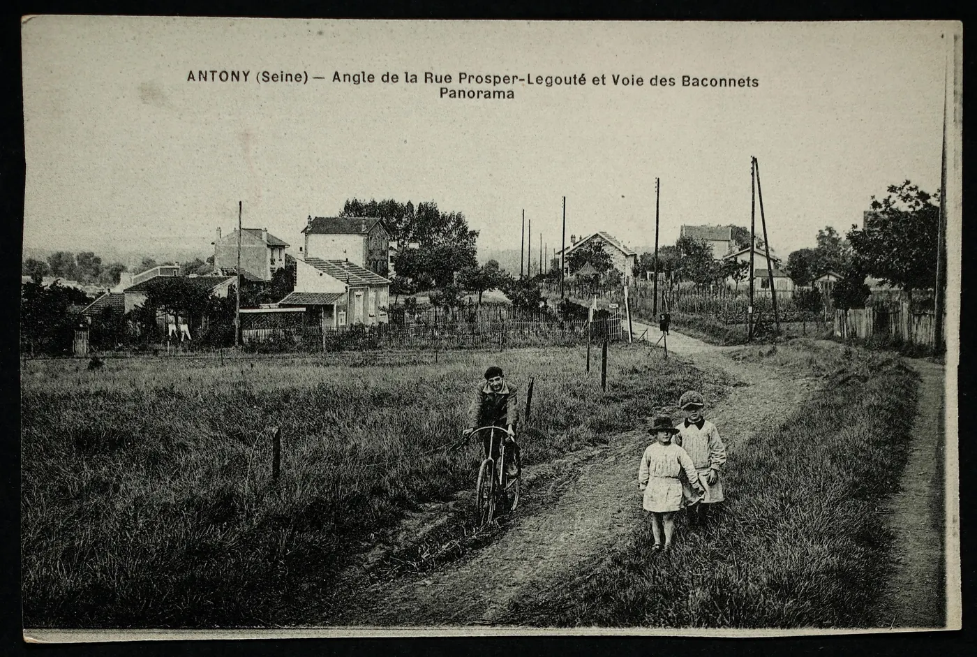 Carte postale ancienne « Antony (Seine), angle de la rue Prosper-Legouté et voie des Baconnets, panorama » : chemins de terre dans un pré, un garçon à vélo et deux enfants, quelques maisons au loin.