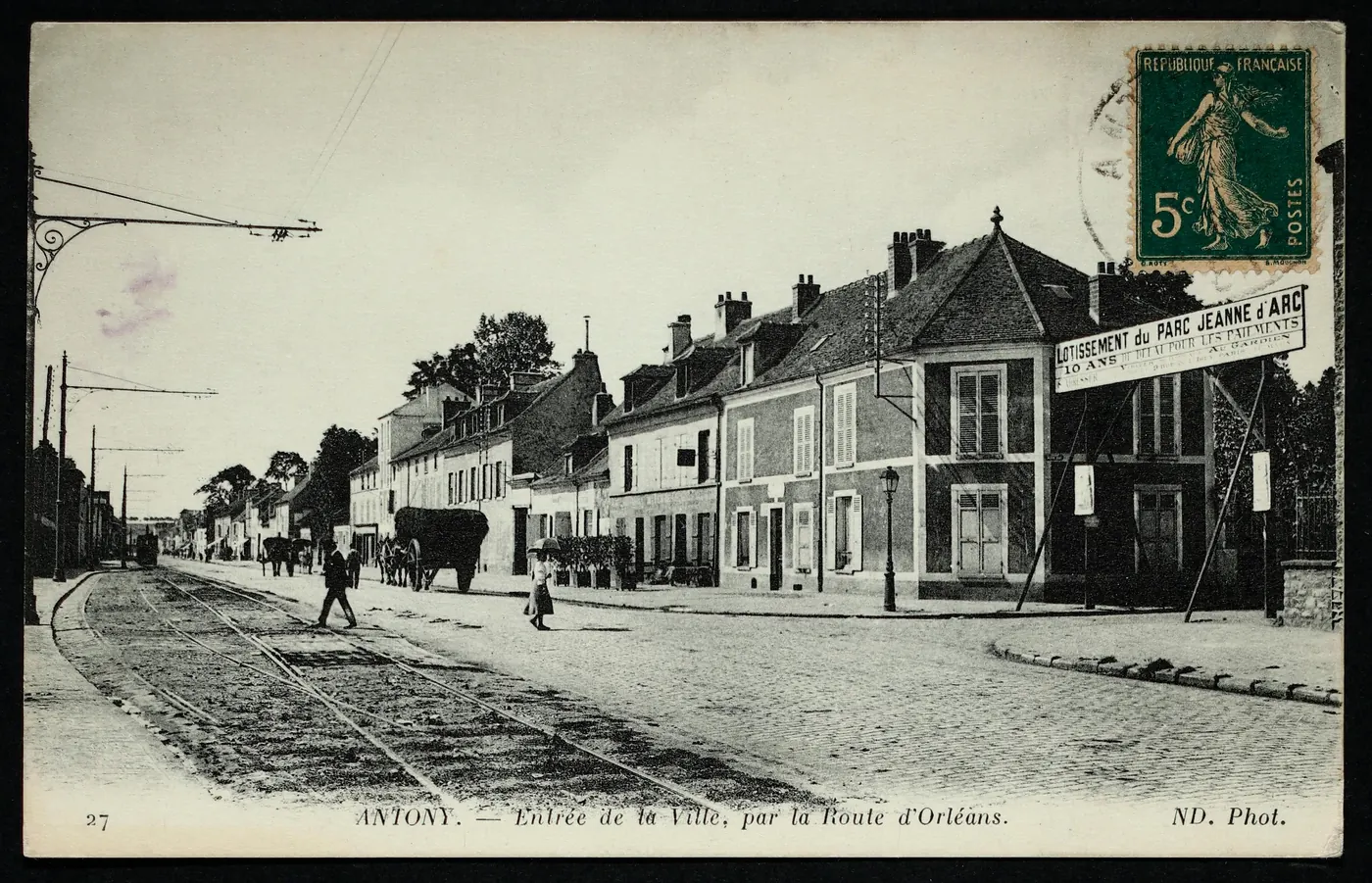 Carte postale ancienne « Antony, entrée de la ville par la route d'Orléans » : rails de tramway, charrette, passants, maisons basses et panneau du lotissement du parc Jeanne d'Arc.
